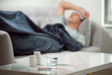 Fotobehang sick wasted man lying in sofa suffering cold and winter flu virus having medicine tablets in health care concept looking temperature on thermometer © makistock