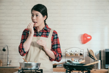 female in pinafore standing in modern wooden kitchen eating piece of chocolate before melting in...