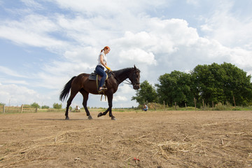 A redhead woman riding a horse on the field. A green bush on a background
