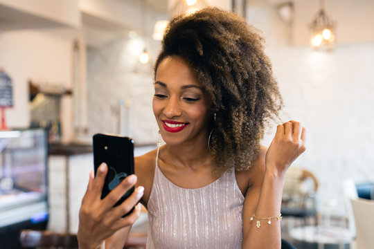 Stylish happy woman showing afro hairstyle taking a selfie photo with smartphone in a cafe bar .