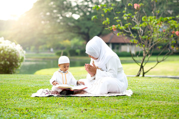 Muslim mother teaching her little son to reading the Quran on grass field near beautiful lake....