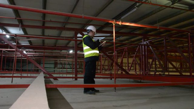 Technician engineer in a helmet checks the sprinklers pipes and roof constructions with a tablet