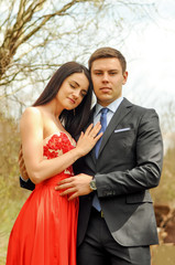 Vertical portrait of the young beautiful, laughing, romantic man and woman wearing long red dress who are looking to the camera outdoors in park in spring or summer