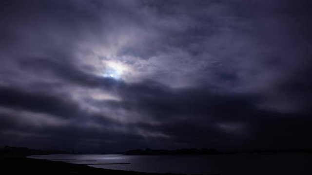 Time lapse at night over a lake with moving cloud fields and light beams from the moon