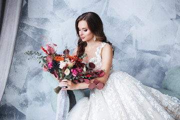 Wedding. Bride in beautiful dress sitting on sofa indoors in white studio interior like at home. Attractive caucasian brunette model like a bride