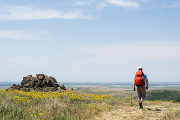 Hiker with a red backpack on a trail with yellow flowers, in Macin Mountains, oldest mountains in Romania, passing near rocks with a small cross on top of them, during a sunny day with clear skies.