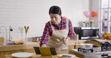 Beautiful girl in apron cooking in kitchen standing near stove. young woman using digital tablet watching screen touching looking at online recipe process. female handmade chocolate on valentine