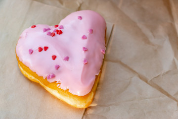 Pink heart donut over the brown paper bag on the wooden table. The closeup food photo.