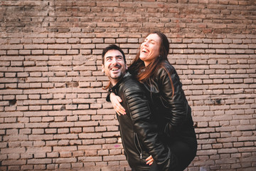 Fototapeta na wymiar Young girl climb on the back of her boyfriend, piggyback, in front of a brick wall having fun and laughing loudly.