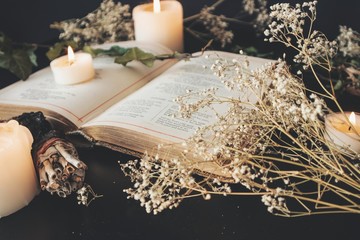 Fototapeta na wymiar Close up of open vintage poetry book decorated with dried baby's breath flowers. Blurred background with white lit burning candles, plants and small sage stick. Black table surface. Romantic soft feel