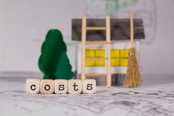 Word COSTS composed of wooden letter. Small paper house, wooden trees in the background.