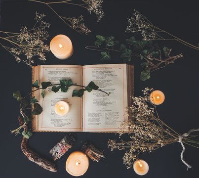 A flat lay of an open vintage book on black table surface with white lit burning candles and dried flowers. Dark romantic cozy feel with a branch of ivy on top of the pages. Square image photo