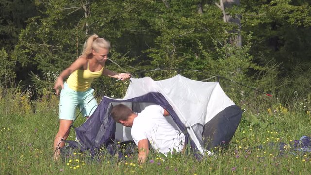 Funny shot of young couple arguing while setting up their tent on a sunny day. Annoyed girlfriend argues with her boyfriend while they set up their campsite by the forest. Young newlyweds fighting.
