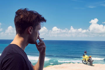 Young handsome man smoking cigar while standing near motorcycle on the tropical beach.