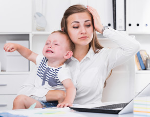 Woman is working with a naughty child in her arms behind a laptop