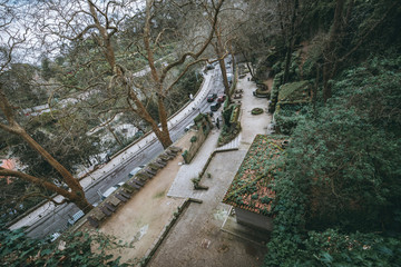 A view from above of the park in Sintra, Portugal, near a bending road with cars: a cracked stony floor, beautiful trees, antique tiled roofs of outbuildings, row of tables with benches; wide-angle