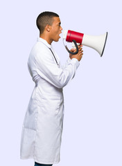 Fototapeta premium Young afro american man doctor shouting through a megaphone to announce something in lateral position on isolated background