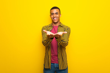 Fototapeta premium Young afro american man on yellow background holding copyspace imaginary on the palm to insert an ad