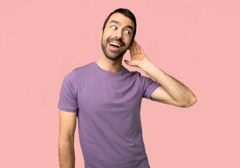 Handsome man listening to something by putting hand on the ear on isolated pink background