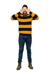 Full-length shot of Handsome man with striped sweater covering eyes by hands. Surprised to see what is ahead on isolated white background