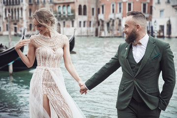 Happy wedding couple near the canal in Venice. Elegant woman in luxury ivory dress, messy updo hair, man in green three-piece suit and bow tie - 249339662
