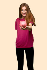 Young redhead girl holding a piggybank on isolated yellow background