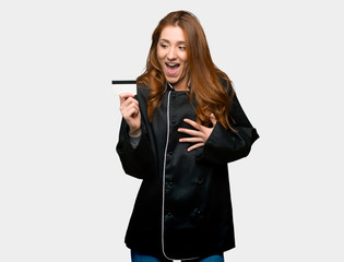 Young redhead chef woman holding a credit card and surprised on isolated grey background