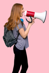 Redhead student woman shouting through a megaphone to announce something in lateral position on isolated pink background