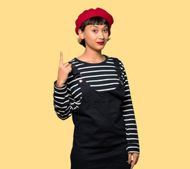 Young woman with beret showing and lifting a finger in sign of the best over yellow background