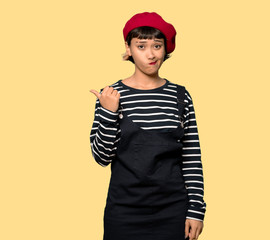 Young woman with beret unhappy and pointing to the side over yellow background