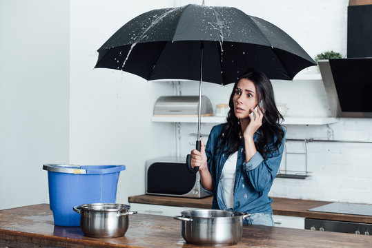 Shocked woman with umbrella dealing with water damage in kitchen and talking on smartphone