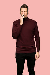 Man with turtleneck sweater yawning and covering wide open mouth with hand over pink background