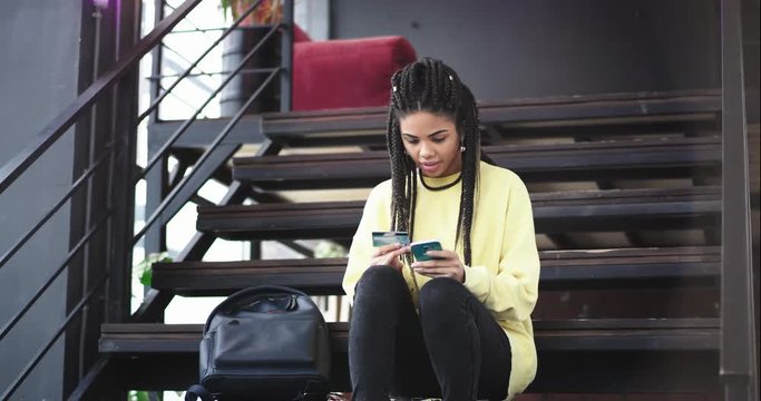 In a modern building with a large window in a sunny day african lady with long hair dreadlocks using a phone and bank card to check something or to buy something online she are very concentrated. 4k