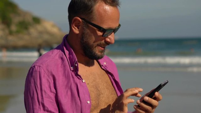 Young, happy man texting on smartphone while standing the on beach
