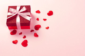 Red gift box with pink ribbon and scattered red hearts on pink background. Christmas, Valentine's day or birthday concept. Place for text.