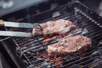 selective focus of juicy raw steaks grilling on barbecue grid with smoke and tweezers
