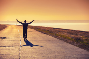 A man with hands in the air walks on the concrete road along the sea at golden sunset