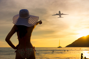 Silhouette of a woman looking to the beach with the sun hat and bikini looking out to sea and the pointing an airplane. Looking landing airplane at Phuket international airport, Thailand