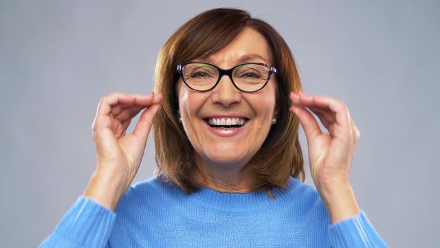 vision, sight and people concept - portrait of happy senior woman putting glasses on over grey background