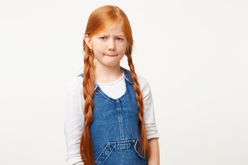 Emotional kid looks sad, displeased, expresses offense,crying, bites her lips, showing defenseless, wears denim overall, stands isolated over white background.