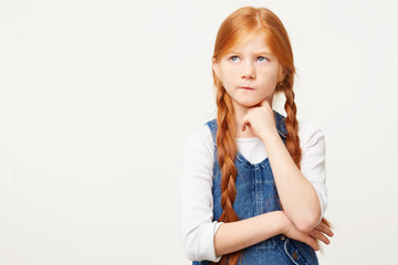 Small red-haired child girl with two long plaits thinks, looks to the left upper corner incredulously with doubt, one corner of the lips pursed, keeps fist under chin,white background,in denim overall