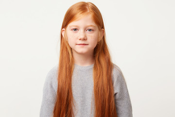 Cutie baby with long red hair down calmly looks into the camera. Beautiful girl behaves with restraint to observe the rules of behavior,over white background