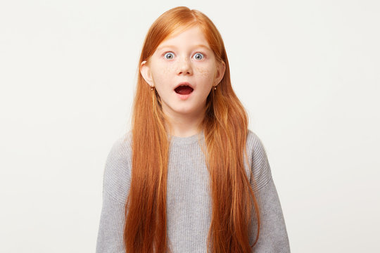 Red-haired girl in shock, nervous frozen in place, stands with her mouth wide open and bulging blue eyes in fear. The child saw something forbidden unexpected scary.