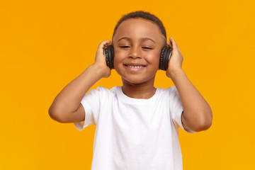 Technology, leisure, music and entertainment concept. Picture of adorable cute African child with broad smile, enjoying favorite songs using wireless headphones. Black little boy listening to music