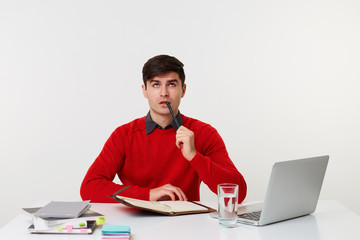 Office manager looking up thinking about his plans, tries to remember something while making up a schedule, keeps pen near lips, sits at the desk on which the laptop books lay, on a white background