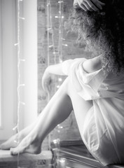 Blurred portrait of a curly girl with bare feet sitting in a men's white shirt by the window. No face, BW photo