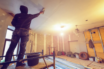Worker is working with palette-knife for flattening the ceiling from wooden platform in room of...