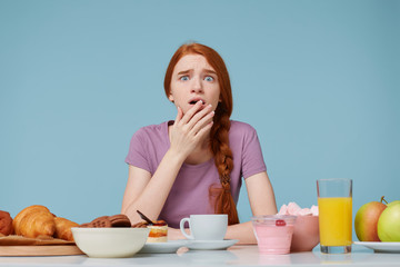 Frightened scared red-haired girl covers open mouth with a palm, sits at a table on which there are many different foods baking fresh fruit,worrying about her health figure, diet violation