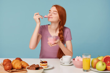 Cute red-haired girl trying tasting cherry yogurt, closed her eyes from pleasure, licks a teaspoon...