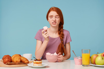 A red-haired girl sitting at a table during breakfast tried a fruit marshmallow, holding a piece in her hands looks at him with disgust, she didn’t like the taste isolated on a blue background.